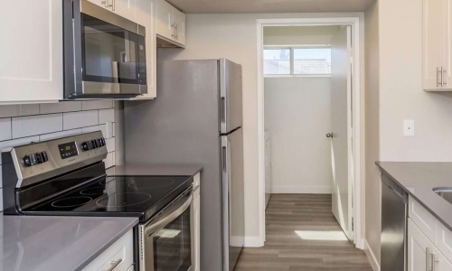 A kitchen with white kitchen cabinetry, a kitchen sink, and appliances including a fridge, microwave, an oven, and a stovetop, with views of a laundry room with a window in a one bedroom apartment at The Solena Apartments in North Las Vegas, NV.